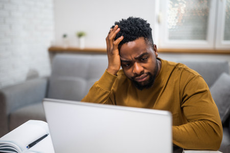 Stressed young African American businessman having a headache while working on laptop at his home.の写真素材