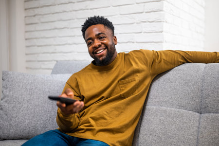 Happy and relaxed young African American man enjoying watching tv while sitting on sofa in his...