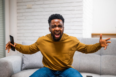 Disappointed young African American man screaming and making angry gestures while watching football game on tv at his home.の写真素材
