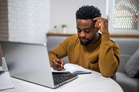 African American businessman sitting at the table, writing notes and using laptop while working at his comfortable home.の写真素材