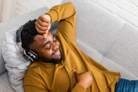 Relaxed young black man with wireless headphones enjoying listening music while lying on sofa in his cozy home.の写真素材