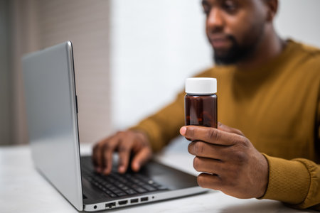 Close up photo of black man holding pill bottle and using laptop for searching more information about his prescription medicine.の写真素材