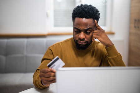 African-American businessman using laptop for online banking, holding his credit card and looking...
