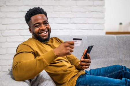 Smiling young African-American man sitting on sofa in modern living room, comfortably using his...