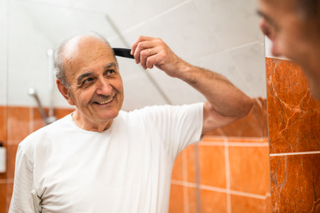 Portrait of happy senior man combing his gray hair while looking himself in the mirror in bathroom.の写真素材