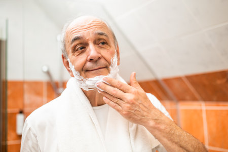Senior man with gray hair looking into bathroom mirror while applying shaving cream.の写真素材