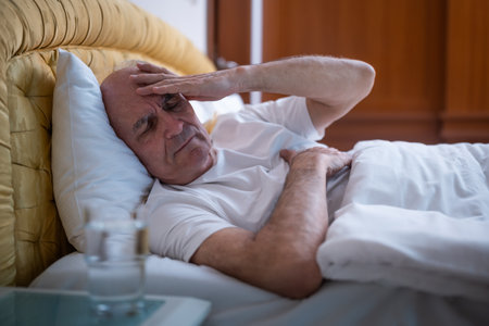 Depressed senior man having a headache while lying in bed in early morning.He is showing signs of mental exhaustion and health concerns in aging process.の写真素材