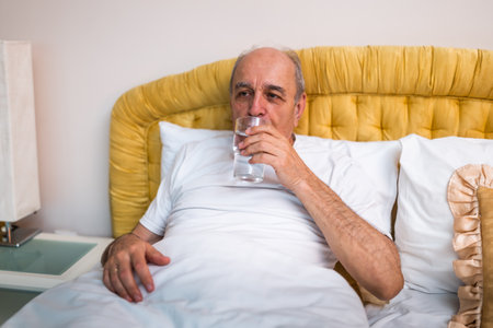 Relaxed senior man drinking water after waking up in bed at his home. The scene reflects contentment, comfort and good health practices in later life.の写真素材