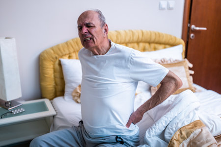 Senior man having pain in back after waking up and sitting on bed in the early morning hours.の写真素材