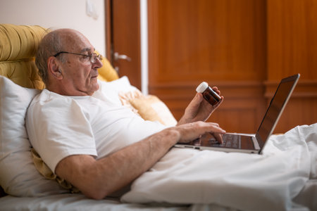 Elderly man in pajamas with eyeglasses searching health information about capsules and pills on laptop while lying in bed at his home.の写真素材