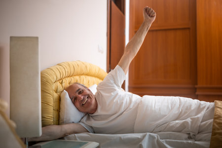 Happy senior man stretching in bed after waking up in morning. The soft morning light and cozy bedroom setting reflect comfort, relaxation and the joys of retirement.の写真素材