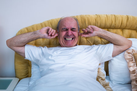 Angry senior man lying in bed and shouting while struggling with noise irritation and insomnia in early morning hours.の写真素材