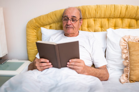 Content senior man with gray hair and eyeglasses reading book in bed, enjoying quiet and cozy morning at home. Leisure and retirement lifestyle.の写真素材