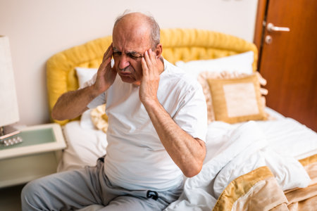 Despaired senior man struggling with headache after waking up in the morning and sitting on bed in softly lit bedroom.の写真素材