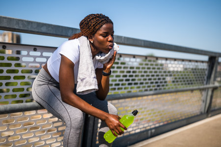Female athlete catching her breath, wiping sweat with towel and drinking sport drink while sitting on a fence of urban city bridge.の写真素材