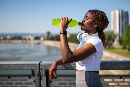 Black female runner pausing her workout outdoor and drinking energy drink to recharge her energy.の写真素材