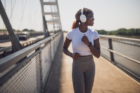African American woman jogging confidently across a city bridge, wearing headphones and sportswear, enjoying her outdoor workout.の写真素材