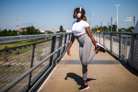 Fit African American woman practicing flexibility exercises in sportswear on urban city bridge.の写真素材