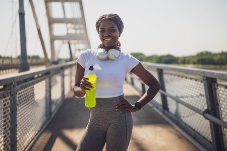Portrait of a happy African American female athlete taking a break from jogging in an urban setting. She refreshes herself with an energy drink, looking energized and motivated.の写真素材