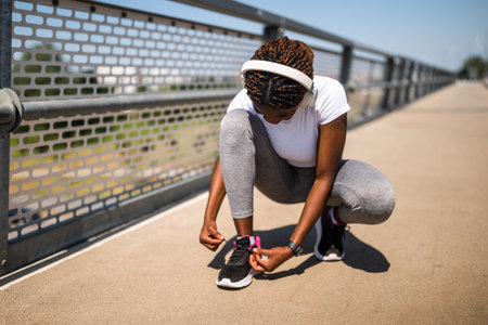 Young sporty African-American woman tying her shoelaces on a city bridge before jogging, wearing headphones and sportswear, ready for workout.の写真素材