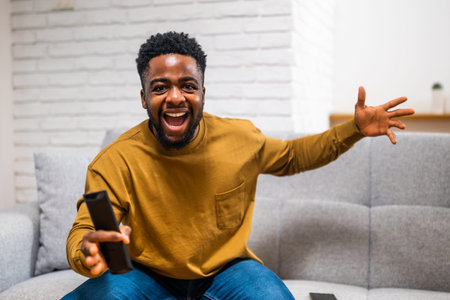 Young African American man making excited gestures while watching football game on tv at his home.