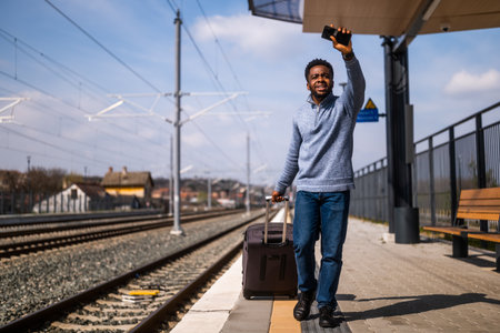 African American man with suitcase shouting and waving at departing train while running along railway station.の写真素材