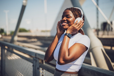 Cheerful African woman enjoying listening to music through headphones after exercise training on urban city bridge.の写真素材