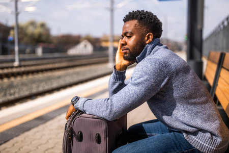 Tired and angry African man with a suitcase sitting on a bench at the railway station.の写真素材