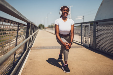 Determined and young black woman in sportswear exercising and stretching while listening to music through wireless headphones on urban city bridge.の写真素材