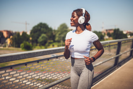 Muscular African - American woman with headphones on jogging outdoors on urban city bridge.の写真素材