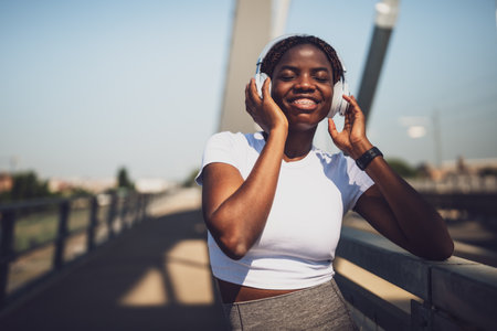 Happy black sporty woman in active wear enjoying listening to music through headphones after exercise training on urban city bridge.の写真素材