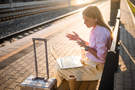 Businesswoman using laptop and earphones while sitting on a bench with luggage at the railway station. She is talking and gesturing during a video call.の写真素材
