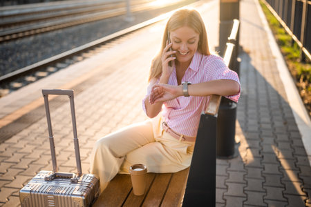 Elegant businesswoman checking the time on her wristwatch and talking on smartphone while sitting on bench with suitcase at the sunny railway station.の写真素材