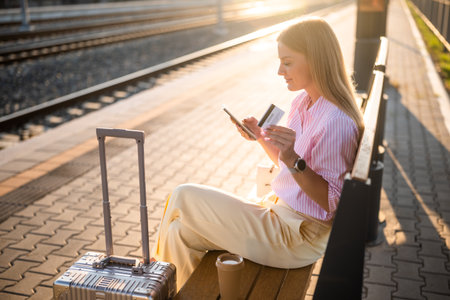 Businesswoman businesswoman using smartphone and credit card while sitting on a bench with suitcase at the railway station and waiting arrival of train.の写真素材
