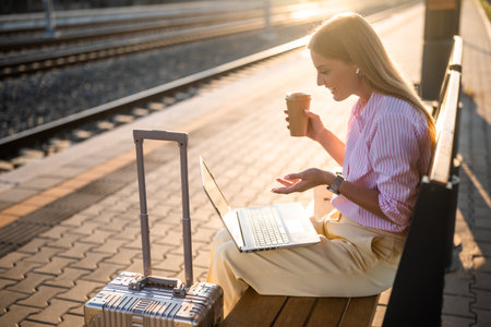 Businesswoman sitting on bench at the railway station platform with suitcase, using laptop and earphones while drinking coffee. She is having a video call.の写真素材