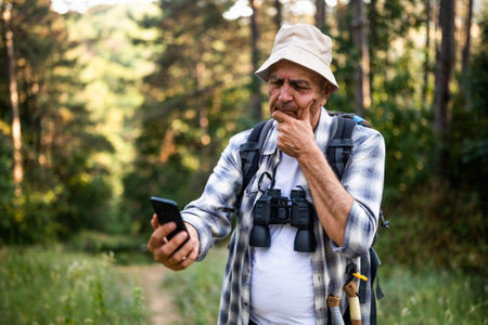 Upset senior hiker got lost and he is using smartphone to find right direction while hiking in the...
