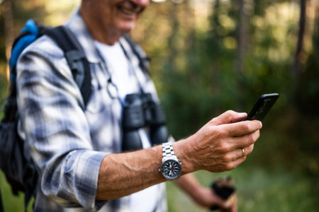 Close up image of senior man with backpack and hiking poles using smartphone while enjoying hiking...