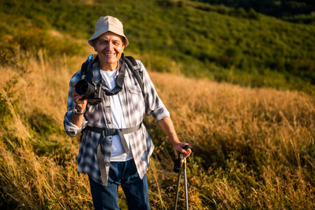 Portrait of happy and active senior hiker using camera for photographing while hiking with backpack...