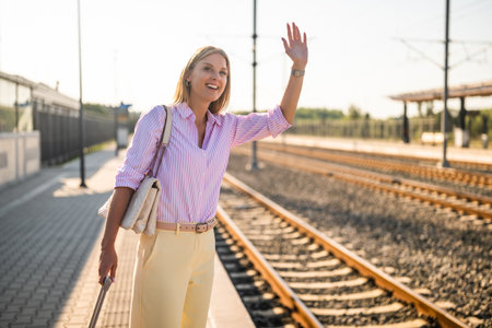 Beautiful businesswoman holding suitcase and waving with a smile while standing on a sunny train platform, dressed in elegant businesswear.の写真素材