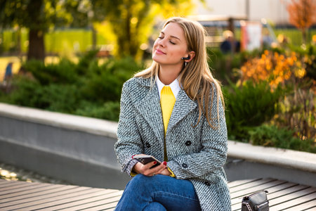 Portrait of beautiful urban businesswoman in autumn coat listening to music through earphones and...