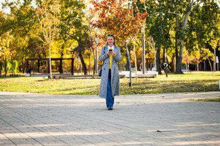 Confident businesswoman with sunglasses in urban coat using smartphone while walking  through the city park during sunny day.の写真素材