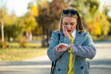 Stressed and worried businesswoman in autumn coat checking the time on her wristwatch while standing in the city park.の写真素材