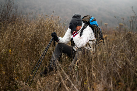 Exhausted and worried hiker in warm clothing sitting alone with backpack and hiking poles in nature...