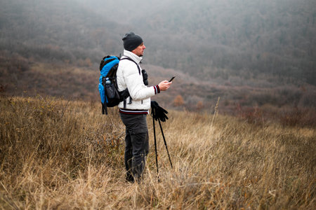 Hiker in warm clothing with backpack and hiking poles using smartphone while enjoying hiking in...