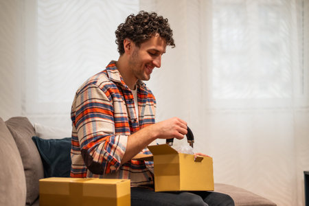 Smiling young man sitting on sofa in living room, opening cardboard box from an online order,...
