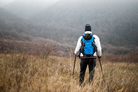 Rear view of hiker in warm clothing standing with backpack and hiking poles standing on a misty...
