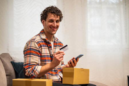 Portrait of smiling young man holding cardboard box and making mobile payment with smartphone and...
