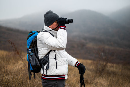 Hiker in warm clothing with backpack and hiking poles using binoculars while hiking in nature on a...