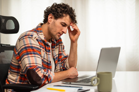 Despaired and exhausted young businessman having a headache while working on laptop at his home.
