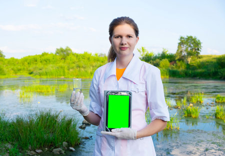 the laboratory assistant stands on the shore of the reservoir with a water sample and a tablet, the...
